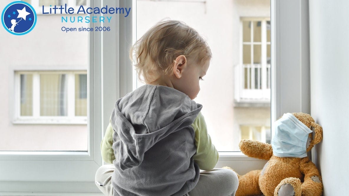 A Baby boy wearing grey shirt facing a teddy bear which is wearing a mask on its face.